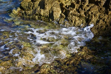 A sea wave crashes against the rocky shore, leaving white foam on the water. The texture of the rocks and the clear water create a feeling of freshness and natural power.