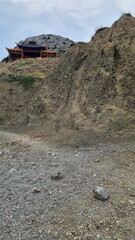 A rocky hillside with a gravel path leading to a wooden structure on top. The sky is cloudy, and...