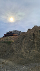 A rocky hillside with a traditional building at the top. The sun is partially obscured by clouds, creating a serene atmosphere.