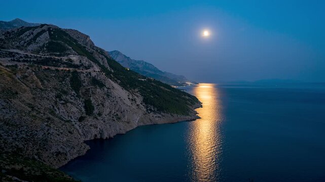 Blue hour timelapse Drasnice Croatia shimmering moon path across Adriatic bay beneath steep Biokovo mountains and coastal highway lights