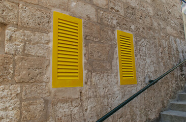 Bold yellow shutters pop against a rustic Maltese stone wall, capturing the charm and contrast of...