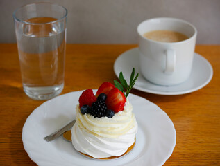 Small pavlova cake with whipped cream and fresh berries served on white porcelain plate with a glass of water and coffee cup on wooden table, minimal and natural light food styling