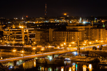 Fototapeta premium Night cityscape of Prague from Letná, Czech Republic, with orange full moon, illuminated skyline and reflections on the Vltava River.