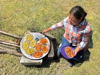 Traditional Mexican woman preparing sopes with salsa on open fire, rustic outdoor cooking of cactus...