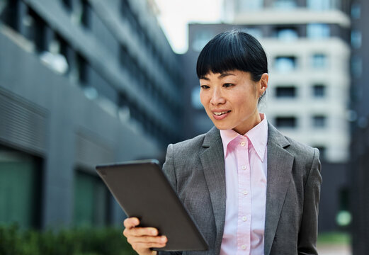 Portrait of a young asian japanese businesswoman woman using a tablet computer and holding a cup of coffee  in a city park, surrounded by modern corporate office buildings