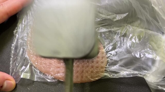 A hand pounds chicken breast under plastic wrap using a metal meat tenderizer on a kitchen counter
