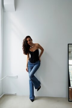 Beautiful tall thin brunette with curly hair in living room. The girl in black korset and jeans, room background. Studio photo shoot for a model. Portrait.