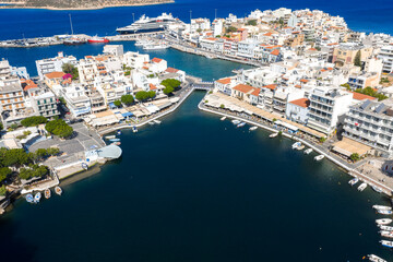 Aerial view of a coastal town in Crete, Greece, with a vibrant harbor, whitewashed buildings, red tiled roofs, yachts, and deep blue waters. © Aerial Film Studio
