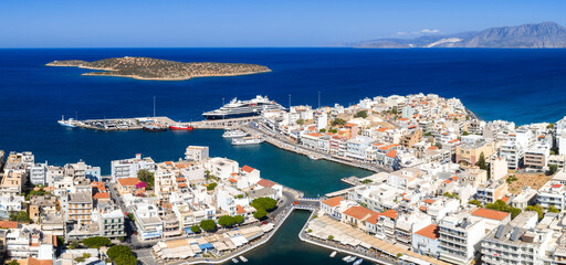 Aerial view of a coastal town in Crete, Greece, featuring whitewashed buildings, a vibrant marina with yachts, and a small island in the Mediterranean Sea. © Aerial Film Studio