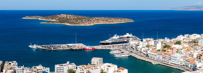 Aerial view of Agios Nikolaos, Crete, showing a harbor with a cruise ship, smaller boats, whitewashed buildings, and a nearby island in the Mediterranean. © Aerial Film Studio