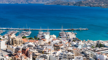 Aerial view of Agios Nikolaos, Crete, showing whitewashed buildings, a domed church, a marina with yachts, turquoise waters, and rugged coastline. © Aerial Film Studio