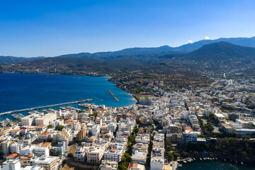 Aerial view of Agios Nikolaos, Crete, showing whitewashed buildings, a marina, a small harbor, the Mediterranean Sea, and rugged mountains. © Aerial Film Studio