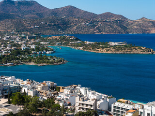 Aerial view of Agios Nikolaos, Crete, Greece, featuring a blue bay, whitewashed buildings, rugged...