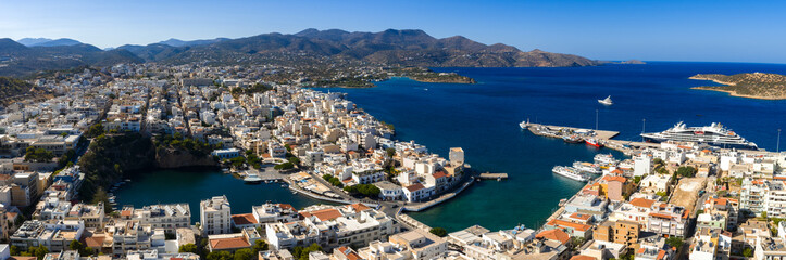 Aerial view of Agios Nikolaos, Crete, showing Lake Voulismeni, the harbor with yachts, white and pastel buildings, and surrounding hills and mountains.