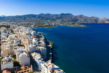 Aerial view of Agios Nikolaos, Crete, Greece, featuring white and pastel buildings, a domed church, a curved bay, and rugged mountains under clear skies.