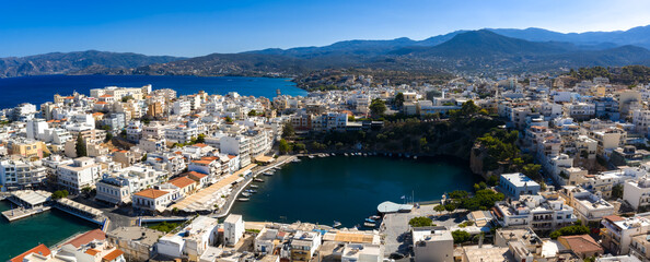 Aerial view of Agios Nikolaos, Crete, featuring Voulismeni Lake, whitewashed buildings, a vibrant waterfront, and the Mediterranean Sea in the background. © Aerial Film Studio