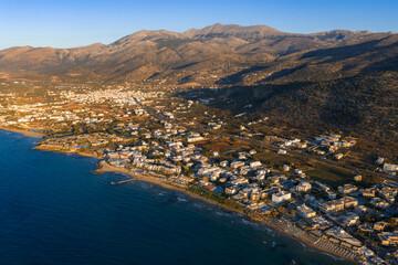 Aerial view of Stalida, Crete, showing sandy beaches, whitewashed buildings, the Aegean Sea, rugged mountains, and surrounding greenery at sunrise or sunset. © Aerial Film Studio