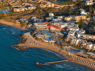 Aerial view of Stalida, Crete, showing a sandy beach with sun loungers, a pier, whitewashed buildings, swimming pools, and turquoise Mediterranean waters. © Aerial Film Studio