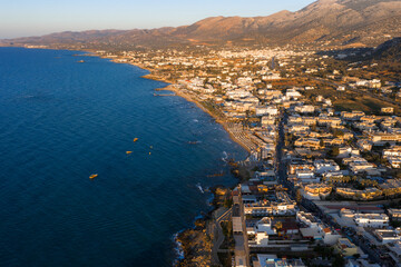 Fototapeta premium Aerial view of Stalida, Crete, showing clear blue waters, sandy beaches, small boats, white and beige buildings, and rugged sunlit mountains.