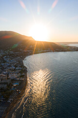 Aerial view of Stalida, Crete, at sunset with golden light over the sea, sandy beaches, whitewashed buildings, and a rugged hill in the background. © Aerial Film Studio