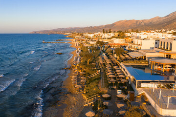 Aerial view of Stalida, Crete, showing sandy beaches with sun loungers, a clear blue sea, a promenade with whitewashed buildings, and rugged mountains. © Aerial Film Studio