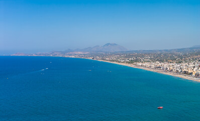 Aerial view of Rethymno town on Crete, Greece, featuring a sandy beach, blue Mediterranean waters, traditional buildings, hills, and a red boat.