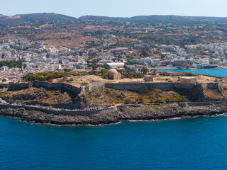 Aerial view of the Fortezza of Rethymno on Crete, Greece, with domed structures, stone walls, the town, rolling hills, and the Mediterranean Sea. © Aerial Film Studio