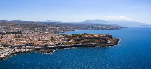 Aerial view of Rethymno town, featuring the Fortezza of Rethymno, terracotta roofed buildings, a Mediterranean coastline, and distant mountains.