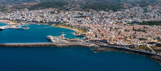 Naklejka premium Aerial view of Rethymno town, Crete, showing white and terracotta roofed buildings, the Venetian harbor with lighthouse, and the Mediterranean coastline.
