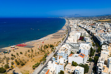 Aerial view of Rethymno, Crete, showing a sandy beach with sunbeds, turquoise sea, whitewashed buildings, red rooftops, and distant mountains. © Aerial Film Studio