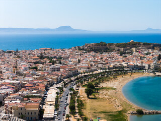 Aerial view of Rethymno town, Crete, featuring the Fortezza fortress, red tiled roofs, a sandy beach, palm lined promenade, and deep blue sea.