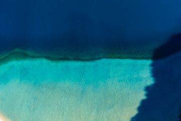 Fototapeta premium Aerial view of Lake Kournas in Crete, Greece, showing a gradient of turquoise to navy blue waters, visible sandy bottom, and contrasting shadows.
