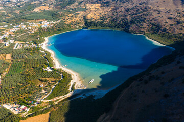 Obraz premium Aerial view of Lake Kournas in Crete, Greece, showing blue waters, sandy beaches, small boats, olive groves, arid hills, and nearby buildings.