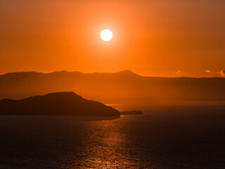 The image shows a sunset over calm waters near Crete Island, Greece, with a glowing horizon, rugged...