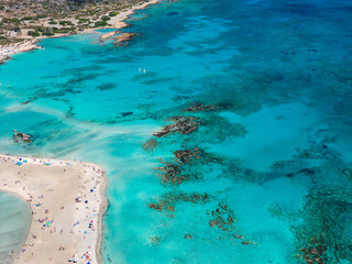 Aerial view of Elafonisi Beach in Crete, Greece, showing turquoise waters, sandy shoreline, sunbathers, umbrellas, and surrounding rugged terrain.