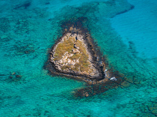 A triangular islet with sparse vegetation and rocky formations surrounded by clear turquoise waters near Elafonisi beach on the island of Crete.