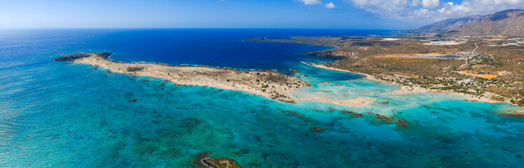 Aerial view of Elafonisi Beach in Crete, Greece, showing turquoise waters, pink sand, shallow lagoons, a sandy peninsula, and rugged mountains.