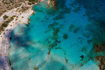 Aerial view of Elafonisi Beach in Crete, Greece, showing turquoise waters, sandy seabed, sun umbrellas, beachgoers, and surrounding rocky terrain.