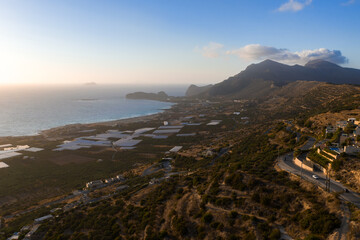 Aerial view of Crete's coastline during sunset, featuring rugged mountains, scattered houses, greenhouses, winding roads, and a tranquil sea.