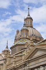 Fototapeta premium Close-up of the ornate dome and colorful tiled roof of the Basilica del Pilar in Zaragoza, featuring intricate spires, sculptural details, and striking Baroque architecture