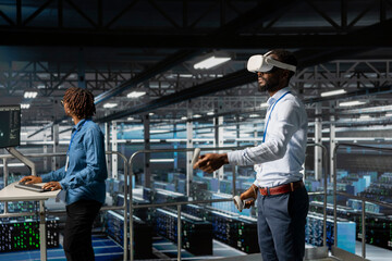 Black male IT expert uses VR glasses on industrial platform in server room, using high tech tools to manage machine learning for automation. Neural network systems in a data center.
