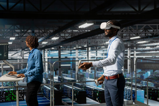 Black male IT expert uses VR glasses on industrial platform in server room, using high tech tools to manage machine learning for automation. Neural network systems in a data center.