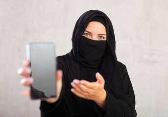young Muslim girl with her face covered by burka shows empty dark smartphone screen. Close-up, gray...