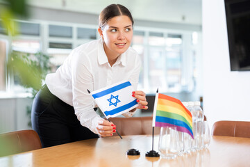 Smiling woman placing flags of Israel and LGBT community on table in office, preparing for...