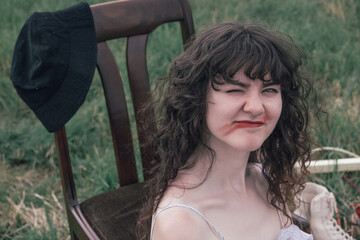 Young woman with messy lipstick winking while sitting on a wooden chair outdoors