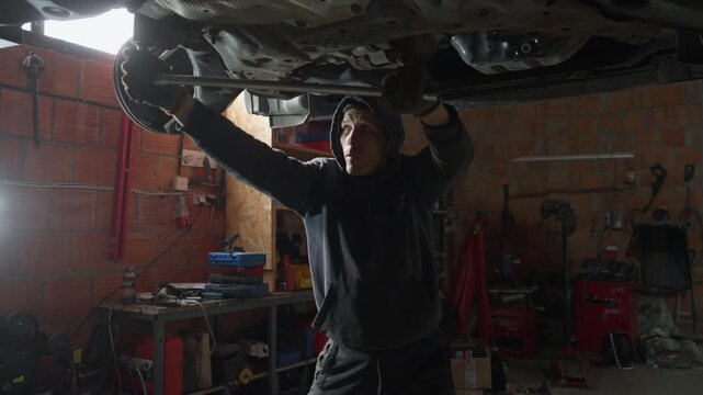 A strong mechanic uses a long wrench for leverage to loosen a stuck bolt on a car. This manual worker is performing heavy-duty vehicle disassembly and maintenance service in a professional garage.