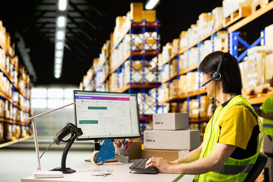 Asian worker in a hi vis vest answers a customer service call in a warehouse logistics center, assisting with tracking shipments, fixing a wrong address issue and updating shipping details.