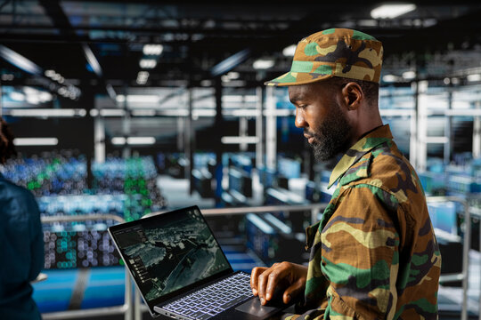 African american military officer in secure data center monitoring AI systems. Armed forces personnel in server farm ensuring stable infrastructure for deep learning defense operations.