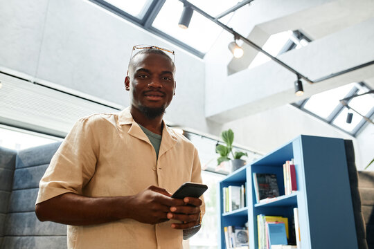 Horizontal medium low angle view shot of handsome young Black man spending free time in modern library holding smartphone looking at camera
