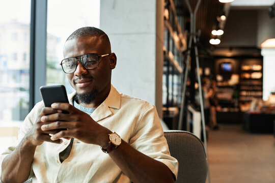 Horizontal medium portrait shot of young African American man with beard on face sitting relaxed in modern cafe watching something on smartphone